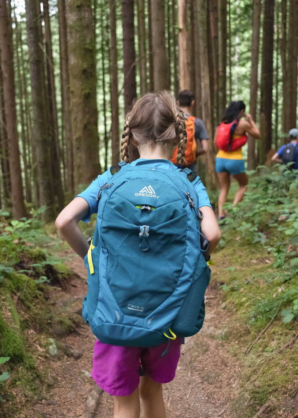 Little girl with two braids in pink shorts, blue t-shirt, carrying a blue Greggory backpack following behind a line of people in the woods